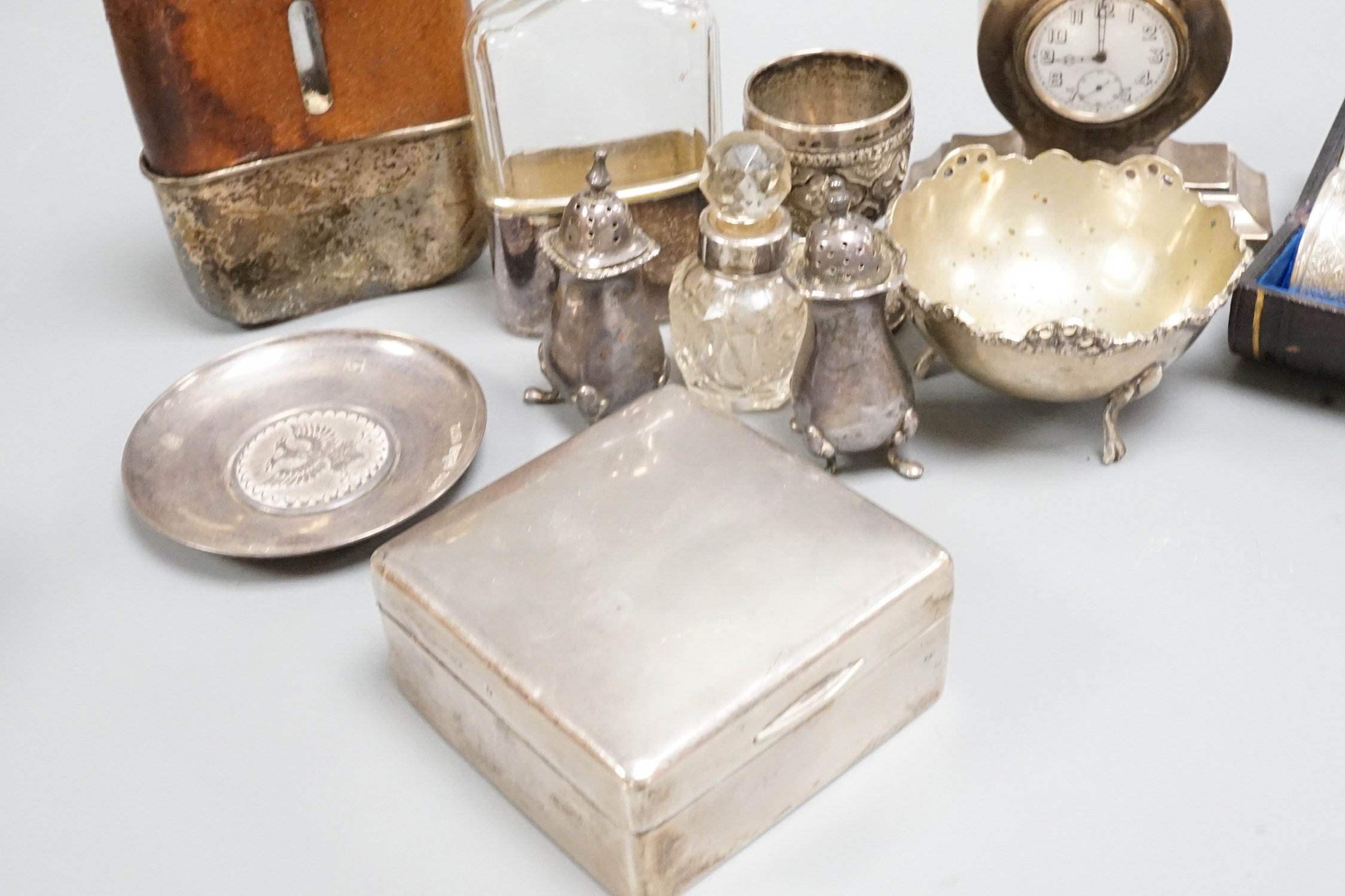 A George V silver mounted small mantel clock, height 93mm, a pair of silver pepperettes and eight other items including a silver mounted cigarette box, a cased set of four chased silver napkin rings and two plated hip fl